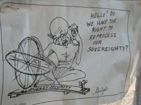 Gandhi with spinning wheel on telephone asking about sovereignty with regards to energy security.  The wheel, a symbol of India’s independence itself is modified to look like the atomic symbol.